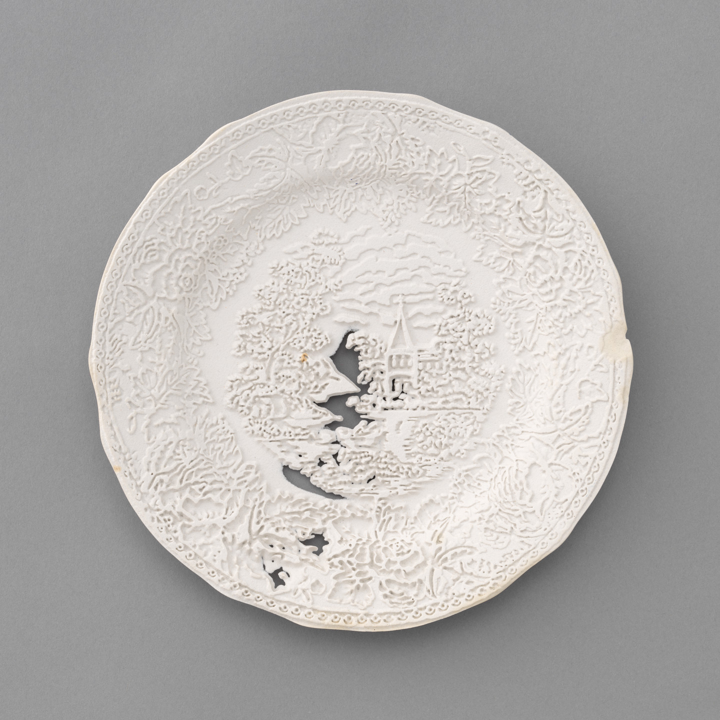 A white, intricately designed ceramic plate features a central scene with trees, a house, a bridge, and a partly cloudy sky, bordered with elaborate floral patterns. The plate has a small chipped section on the right edge. This is an AI generated description. To give feedback, email ai@moma.org.
