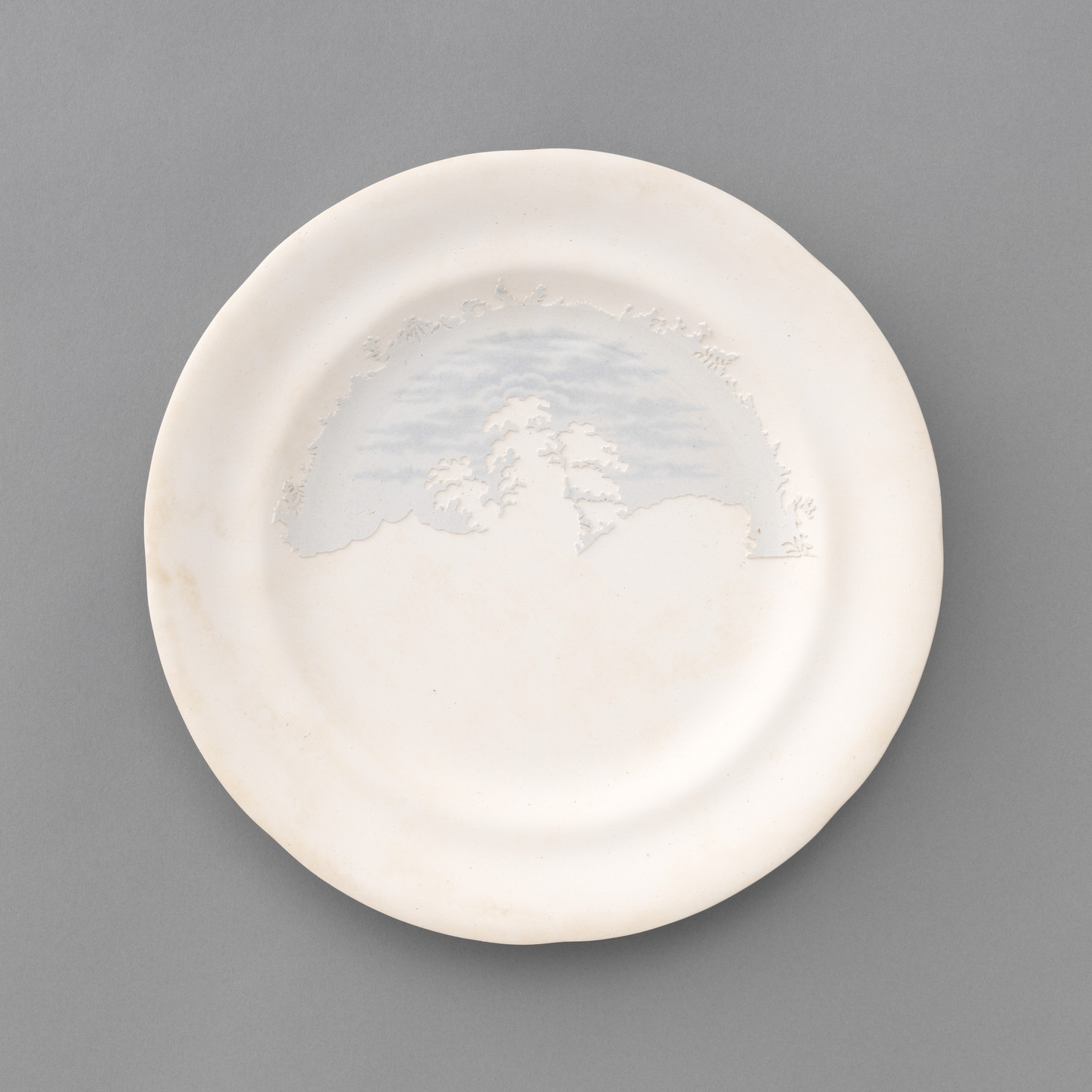 A round, white plate features a subtle, raised depiction of a landscape with trees and cloudy sky in light blue near the upper edge. The plate has a slightly irregular border. This is an AI generated description. To give feedback, email ai@moma.org.