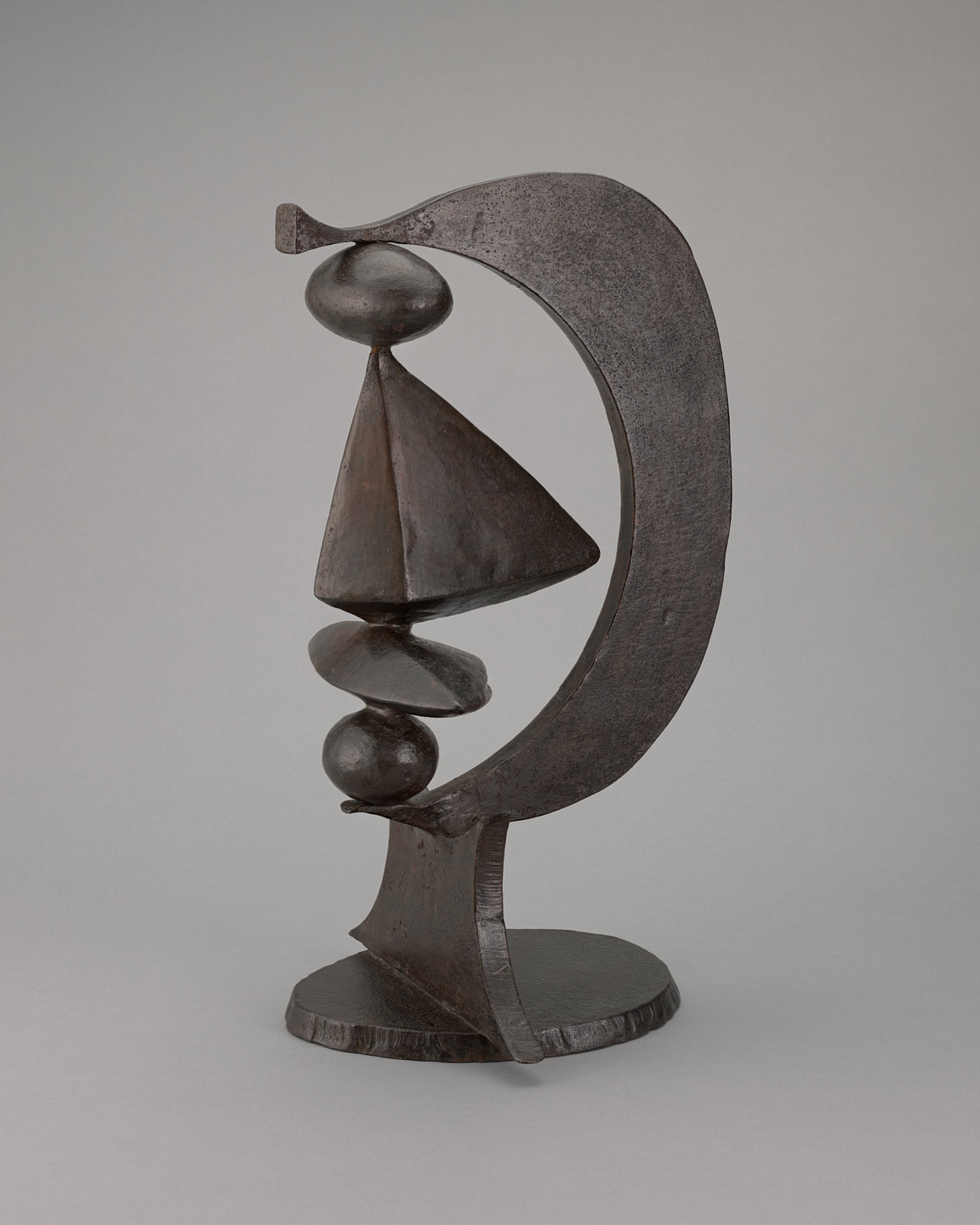 A dark bronze-colored sculpture features geometric shapes, with a cone atop spherical forms, all encircled by a thick, curved arch. The sculpture stands on a round, textured base against a plain gray background. This is an AI generated description. To give feedback, email ai@moma.org.