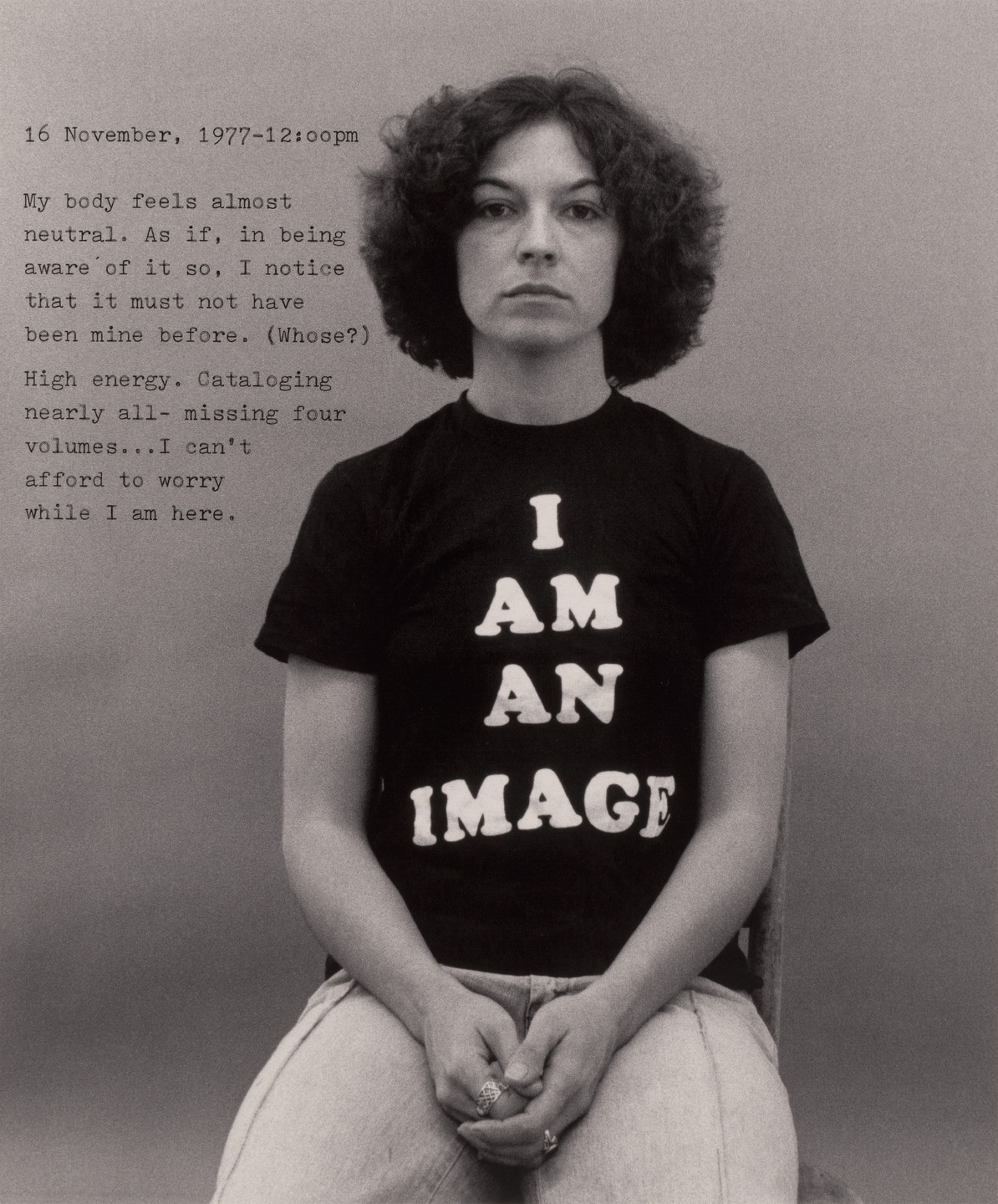 A person with curly hair sits against a plain background, wearing a black t-shirt with &#34;I AM AN IMAGE&#34; in white text. To the left, there is text with a date, time, and a reflection on their body&#39;s neutrality and high energy. This is an AI generated description. To give feedback, email ai@moma.org.