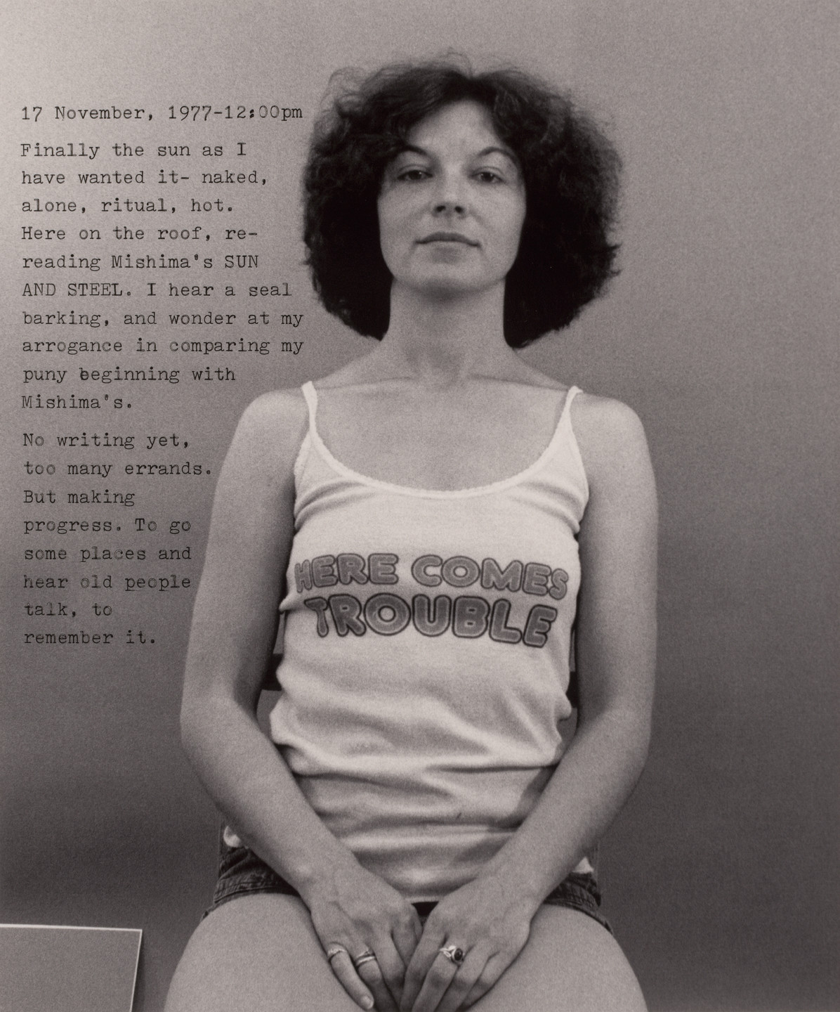 A black-and-white photo shows a person with curly hair in a tank top that reads &#34;HERE COMES TROUBLE.&#34; To the left, text details a reflective entry dated November 17, 1977, at 12:00pm. This is an AI generated description. To give feedback, email ai@moma.org.