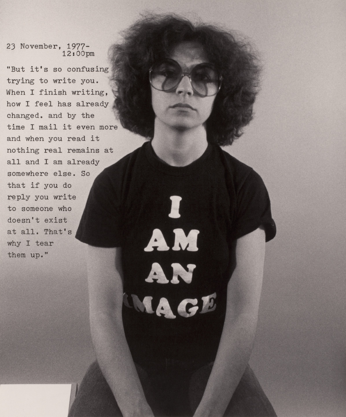 A person with curly hair and large glasses sits against a plain background, wearing a black shirt with &#34;I AM AN IMAGE&#34; printed in large white letters. To the left, a typed note from November 23, 1977, at 12:00 pm is partially visible. This is an AI generated description. To give feedback, email ai@moma.org.