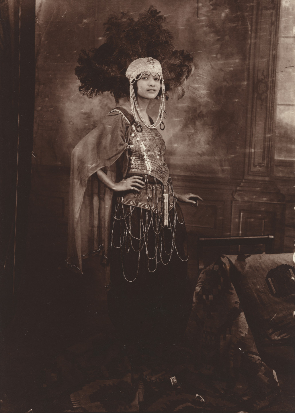A person stands with one arm on hip, wearing a sequin-covered attire with layered chains, a feathered headdress, and a draped sheer fabric. The sepia-toned background shows a decorated wall and a chair draped with fabric. This is an AI generated description. To give feedback, email ai@moma.org.
