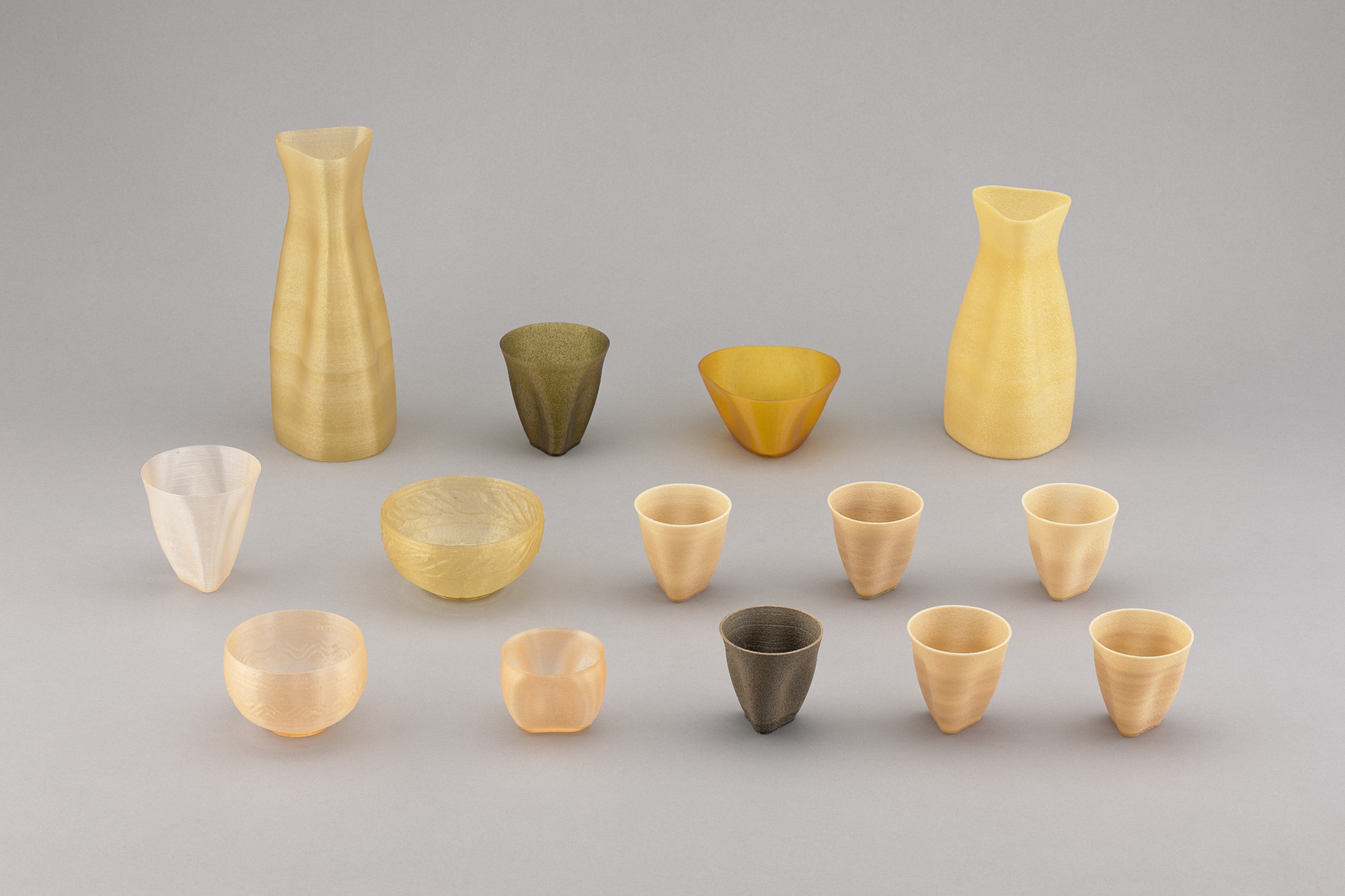 The image shows a collection of 15 vessels, including carafes, bowls, and cups, arranged in three rows on a gray background. Predominant colors are yellow, beige, and brown. This is an AI generated description. To give feedback, email ai@moma.org.