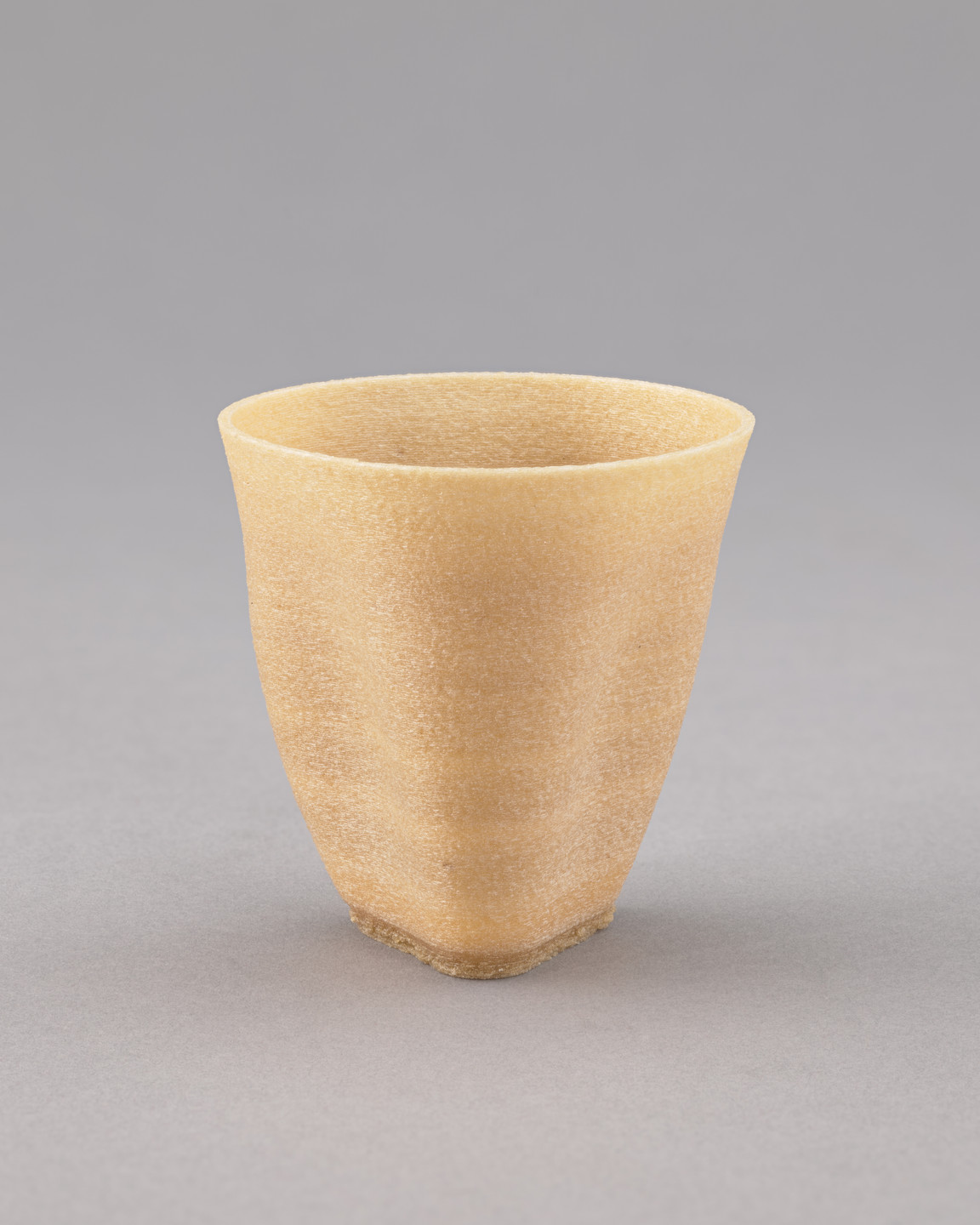 A beige ceramic cup is shown against a plain light grey background. The cup has a textured surface, a slightly flared rim, and a roughly circular shape with a flat base. This is an AI generated description. To give feedback, email ai@moma.org.