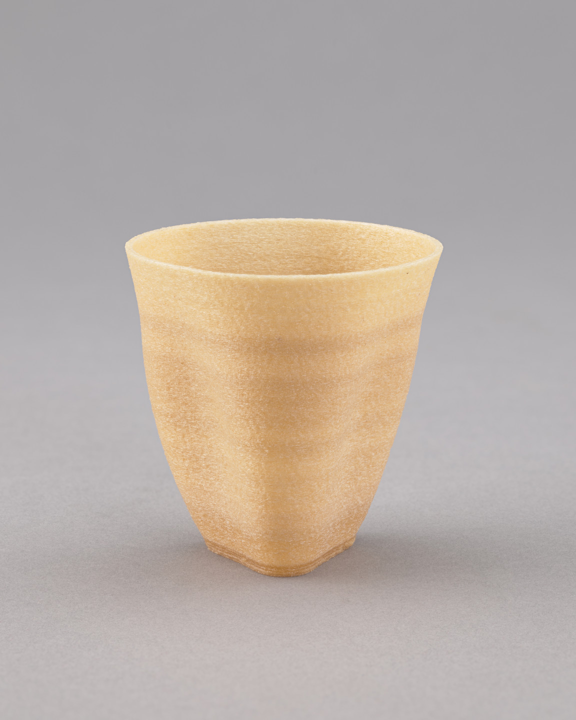 A beige, textured, vase-like object with a slightly flared opening and smooth, vertical contour stands on a neutral gray background. The item has layered horizontal streaks and a matte finish. This is an AI generated description. To give feedback, email ai@moma.org.