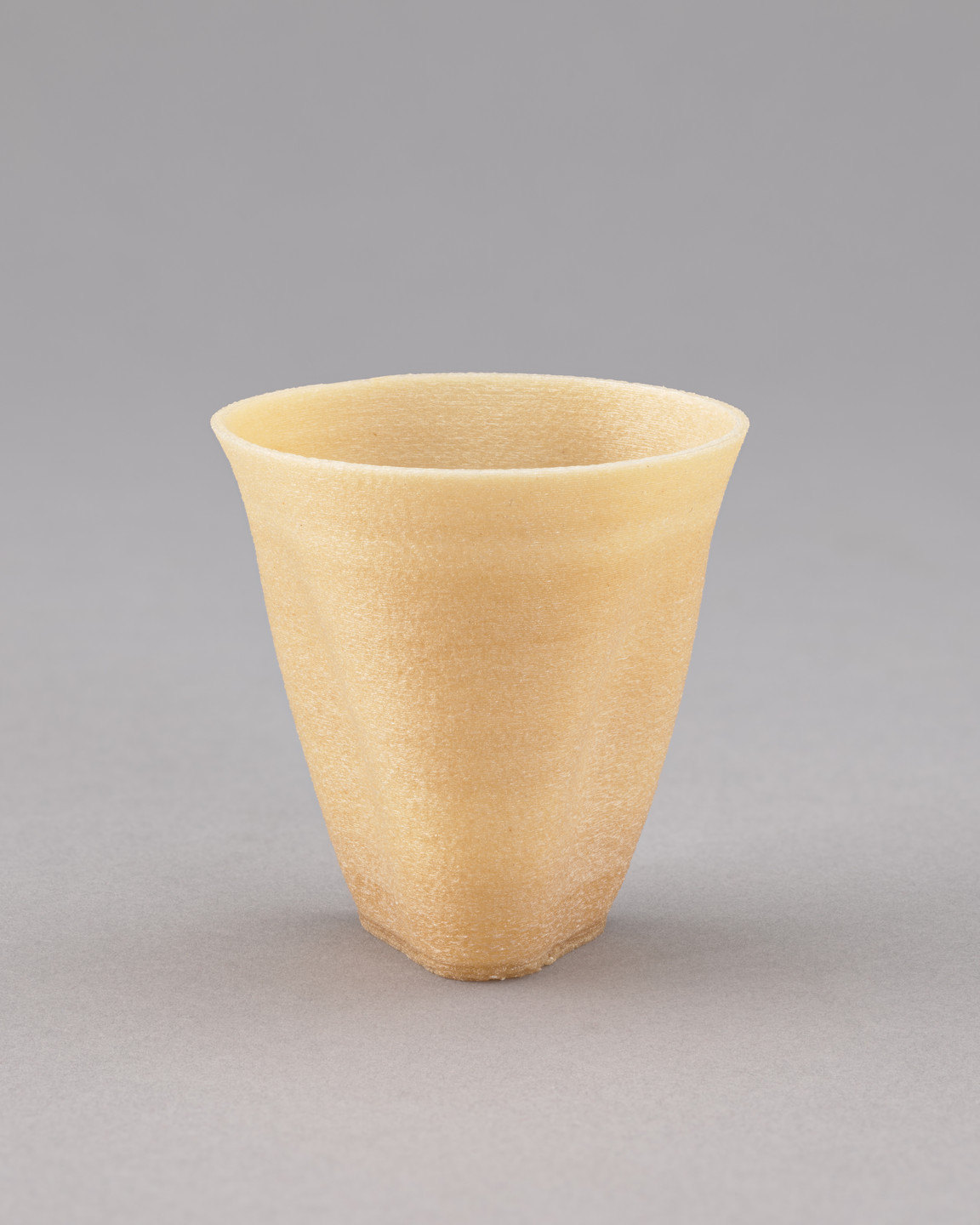 A beige, textured vessel with a wide, open top and a slightly tapering base sits on a light grey surface with a grey background. The vessel appears to be translucent and has a rough surface. This is an AI generated description. To give feedback, email ai@moma.org.