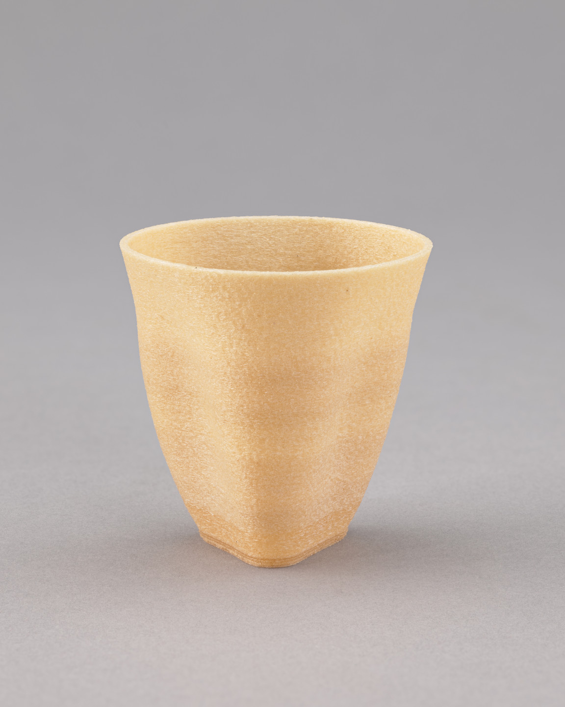 A light brown, textured cup with a slightly flared rim and tapered base is centered against a plain gray background. The cup appears to be made from a rough, sand-like material. This is an AI generated description. To give feedback, email ai@moma.org.