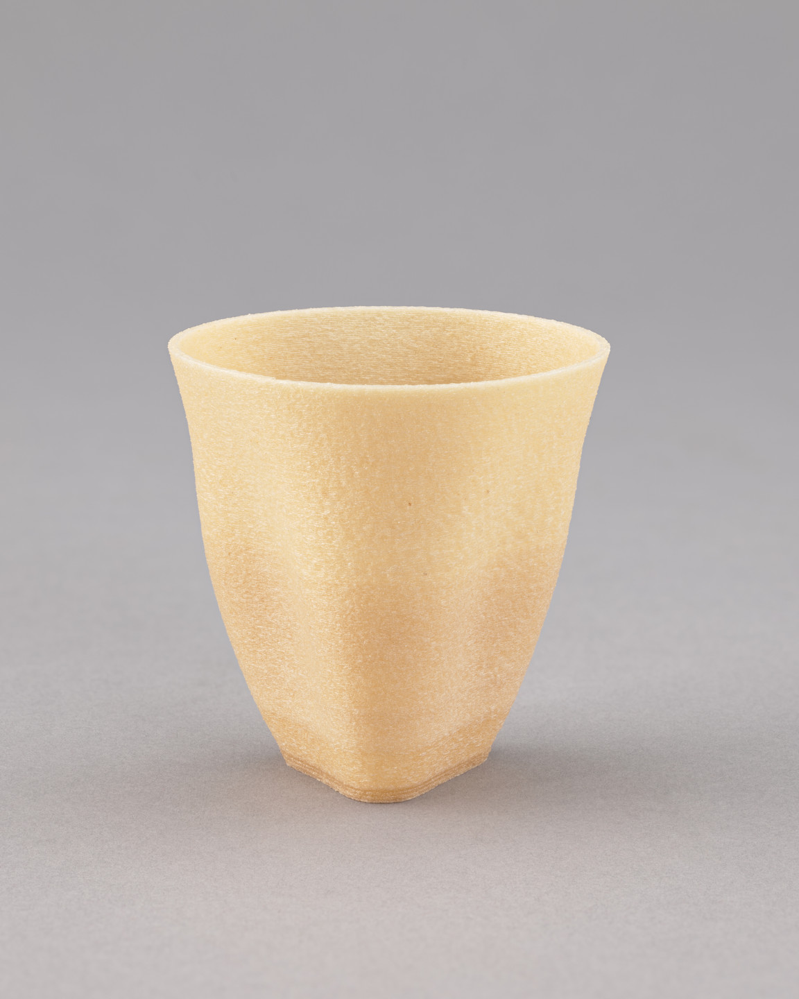 A yellowish-beige cup with a textured surface and smooth, flared top edges is centered against a plain gray background. The cup has a rounded, tapering base and no visible handle. This is an AI generated description. To give feedback, email ai@moma.org.