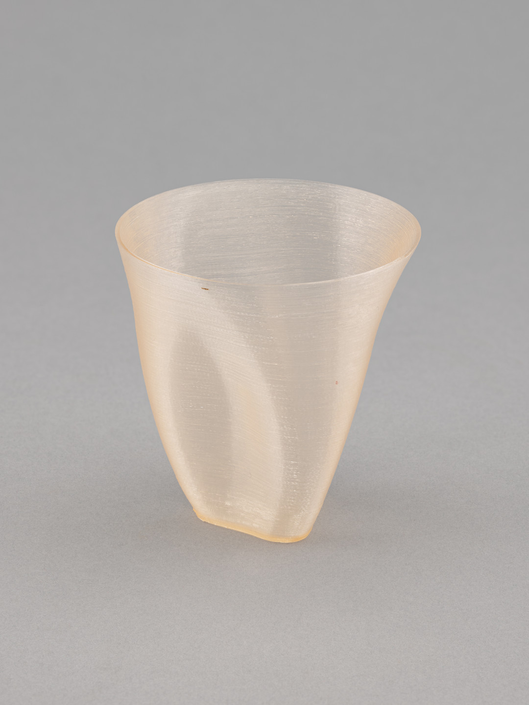A translucent, light beige-colored, slightly flared cup with a smooth, curved surface is set against a plain gray background. The cup has a subtle texture and a tapered bottom. This is an AI generated description. To give feedback, email ai@moma.org.