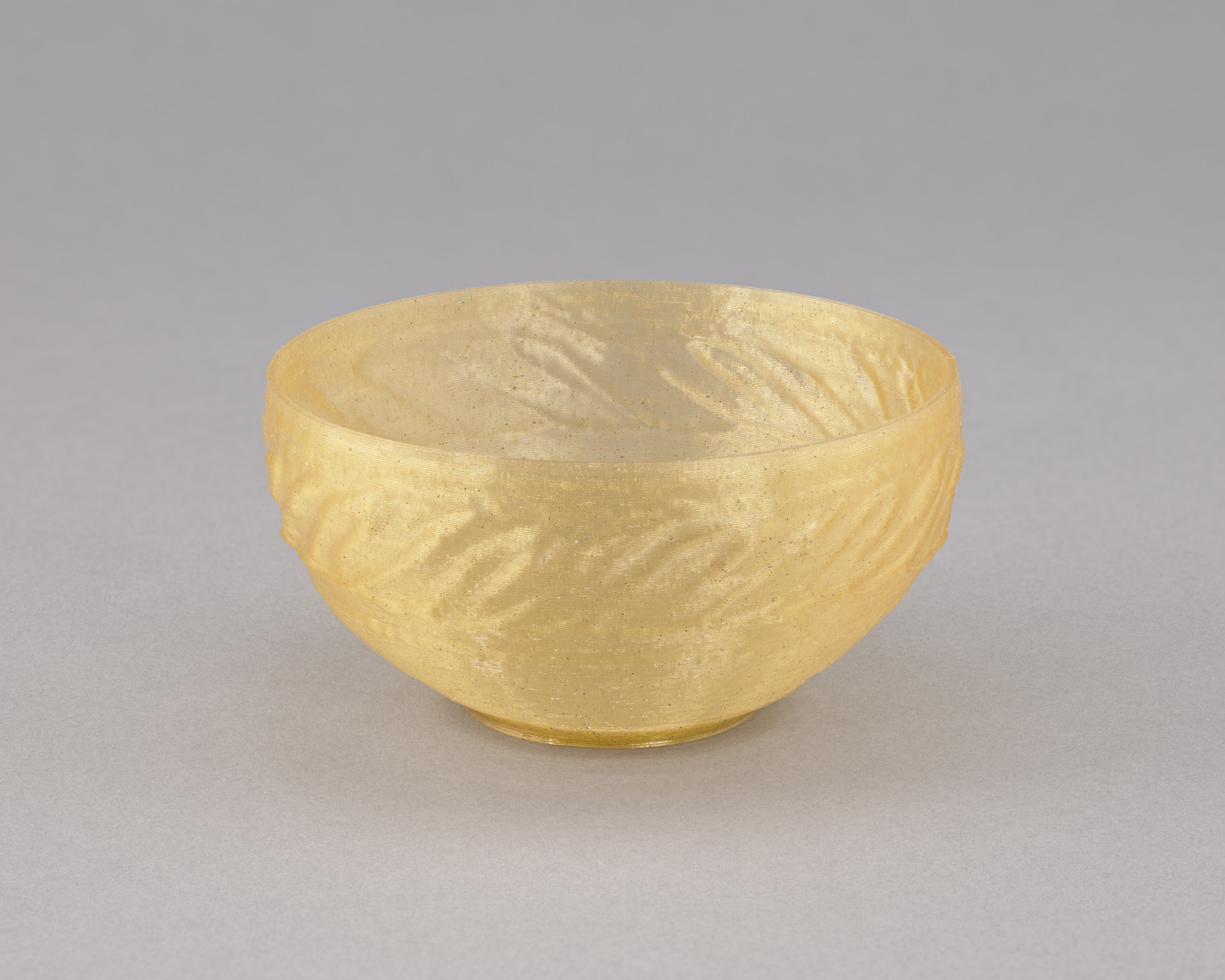A small, transparent bowl with a gold hue and spiraling, textured ridges along its exterior sits on a plain, grey surface. The bowl is empty and appears to have a smooth rim. This is an AI generated description. To give feedback, email ai@moma.org.