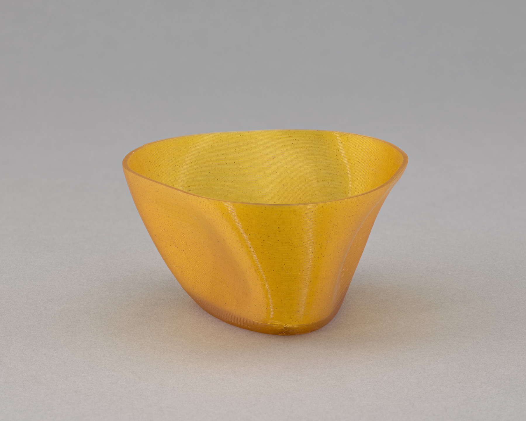 A small, translucent golden-yellow bowl with a smooth, curved surface and an irregular, asymmetrical shape is placed on a plain, light grey background. The bowl has a subtle texture and no visible decorations. This is an AI generated description. To give feedback, email ai@moma.org.
