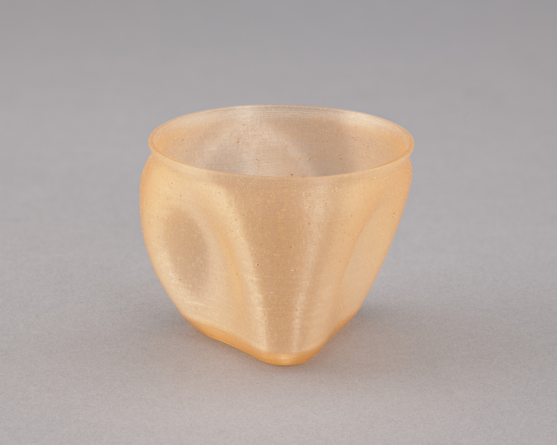 A translucent, peach-colored cup with a smooth, slightly wavy surface sits on a plain grey background. The cup&#39;s shape is somewhat irregular, with a rounded base and slight indentations. This is an AI generated description. To give feedback, email ai@moma.org.