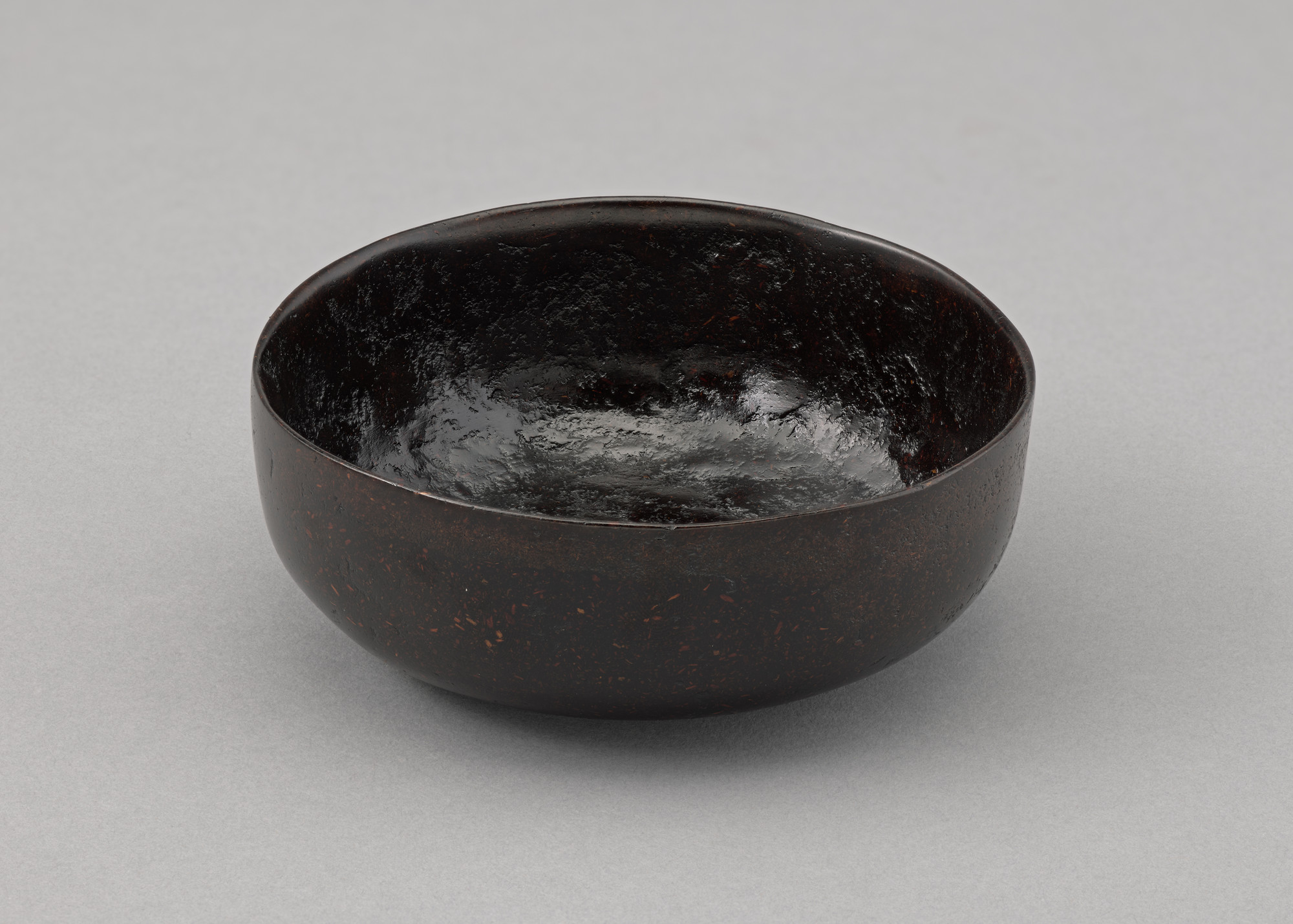 A dark brown, round bowl with a smooth, glossy interior and a slightly textured exterior sits on a light gray surface. The bowl&#39;s rim is uneven, giving it a handmade appearance. This is an AI generated description. To give feedback, email ai@moma.org.