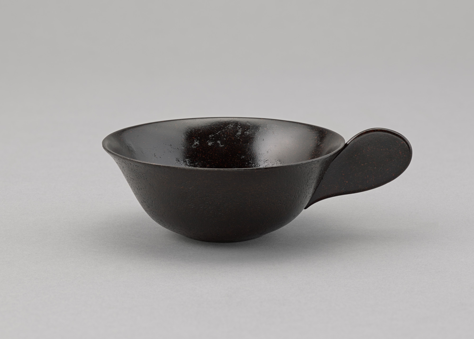 A dark brown cup with a wide, rounded body and a single, thick handle on a light grey background. The cup has a matte, slightly textured exterior and a glossy interior. This is an AI generated description. To give feedback, email ai@moma.org.