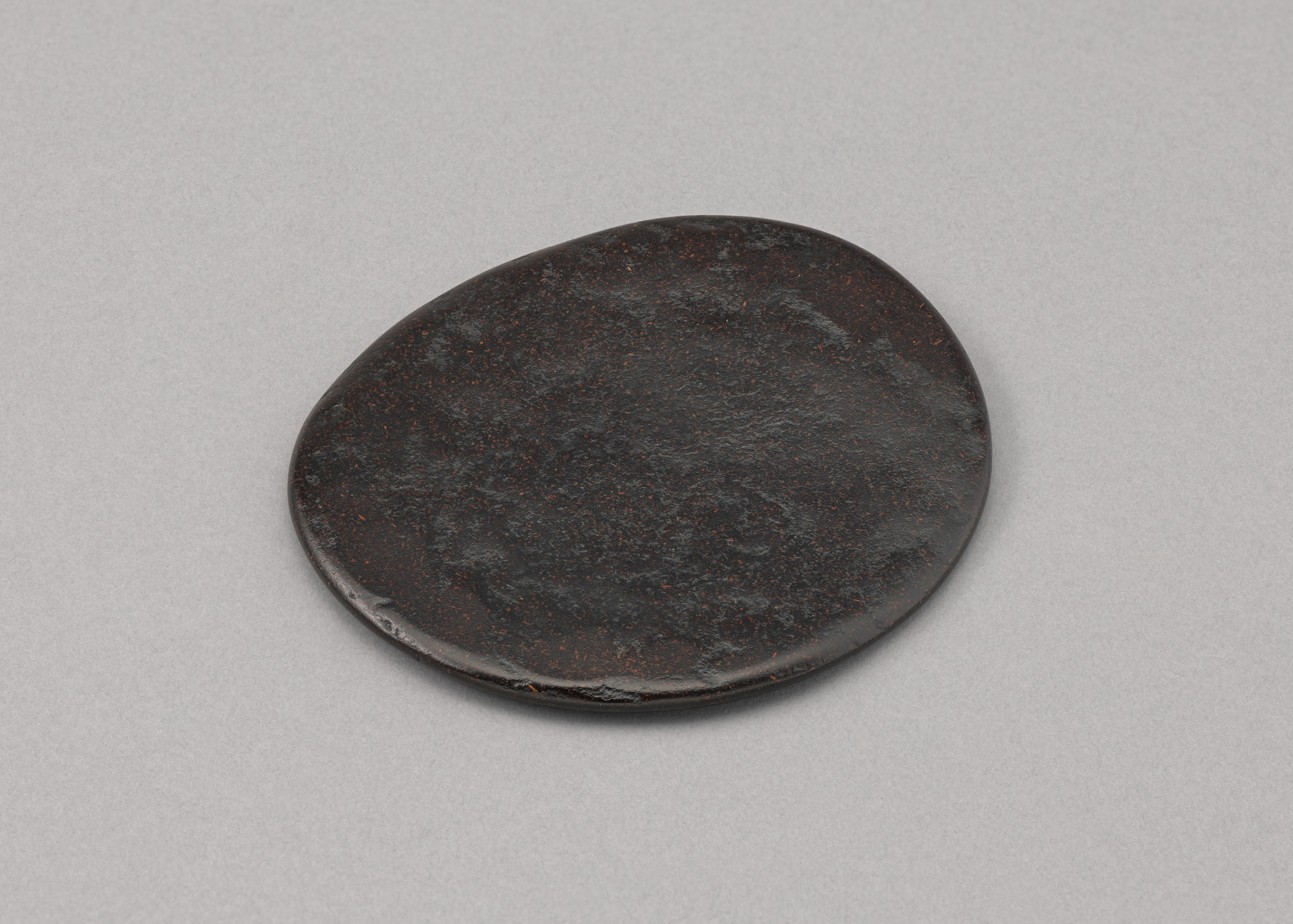 A flat, oval-shaped, dark brown stone rests on a light gray surface. The stone has a slightly uneven texture and appears smooth around the edges. This is an AI generated description. To give feedback, email ai@moma.org.