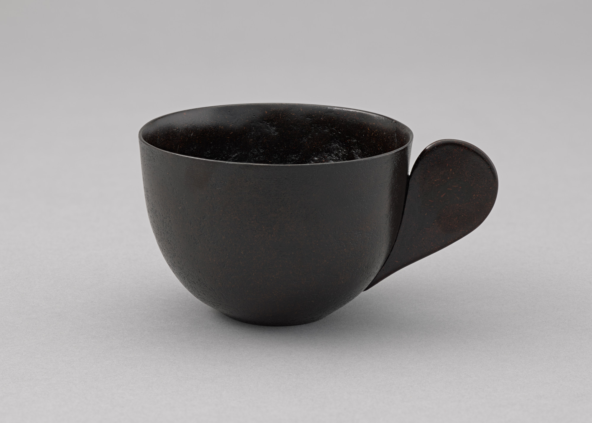 A dark brown cup with a smooth, round body and a large, flat handle sits on a light gray surface. The handle is attached to the side of the cup and curves outwards. This is an AI generated description. To give feedback, email ai@moma.org.