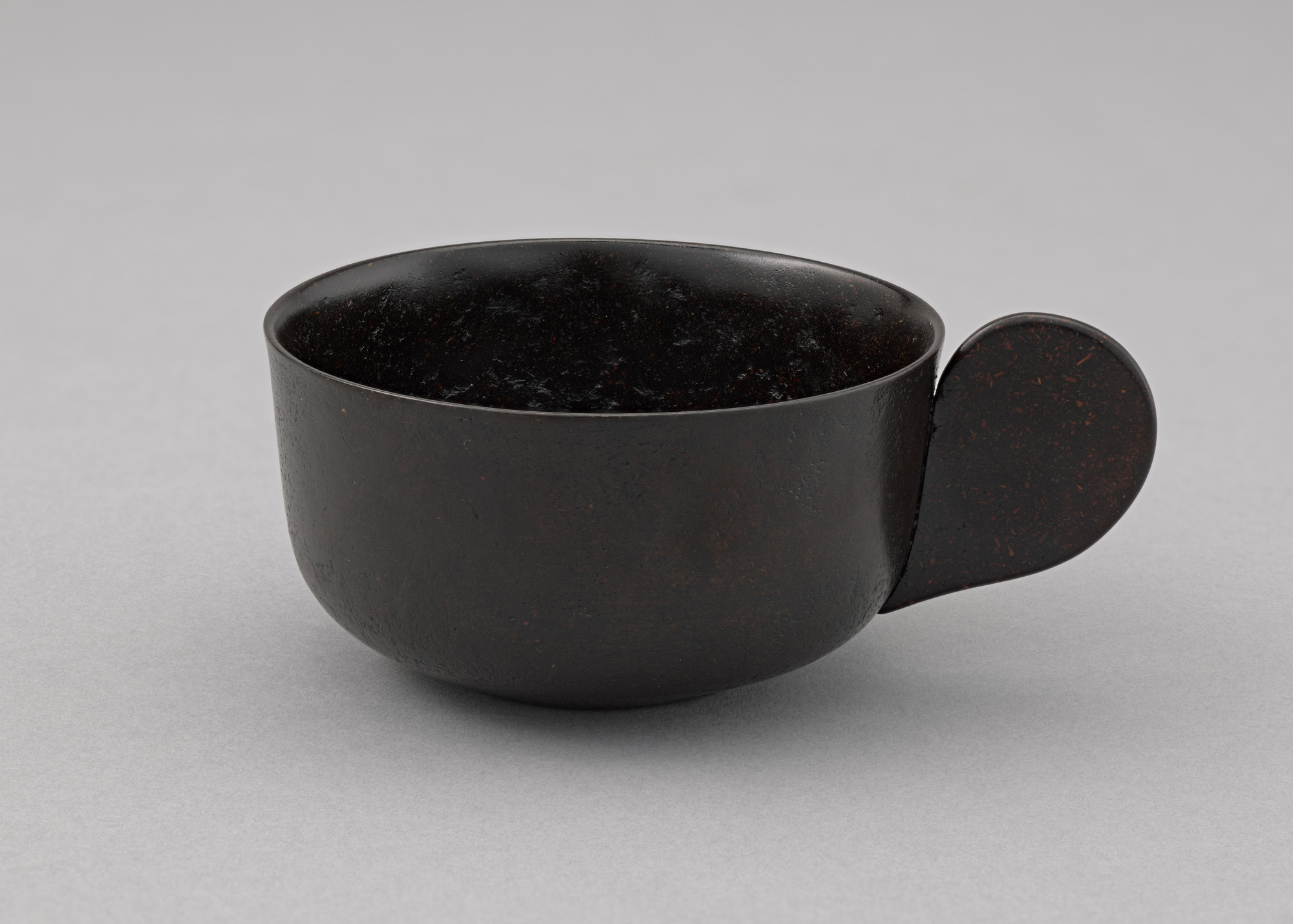 A dark brown bowl with a smooth, round shape sits on a gray background. The bowl features a flat, circular handle extending from one side. This is an AI generated description. To give feedback, email ai@moma.org.
