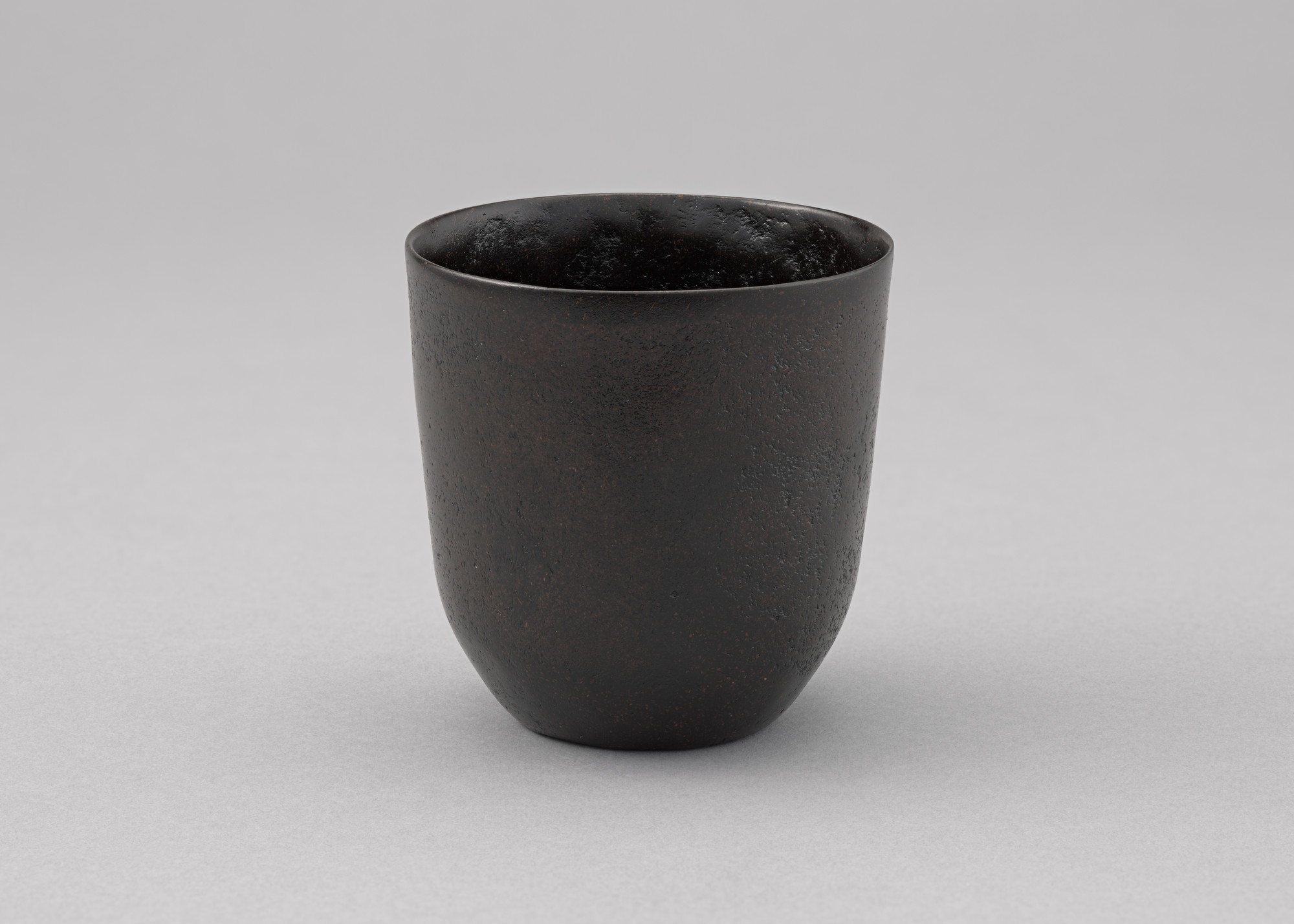 An image shows a dark brown, rustic-looking ceramic cup with a textured surface. The background is a plain, light gray color. This is an AI generated description. To give feedback, email ai@moma.org.