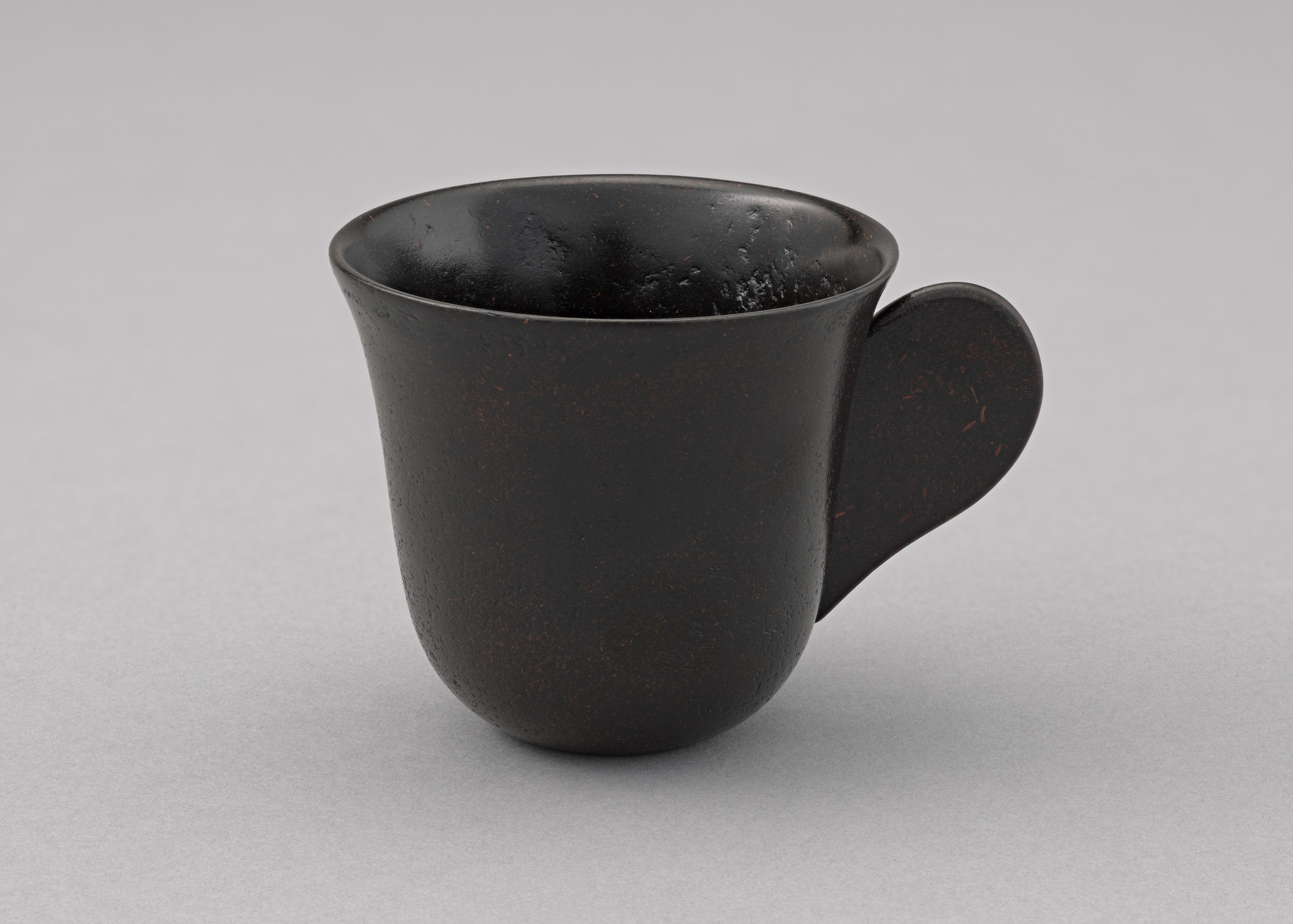 A dark brown, textured ceramic cup with a rounded body and a wide flared rim sits on a gray surface. The cup has a wide, flat handle extending from one side. This is an AI generated description. To give feedback, email ai@moma.org.